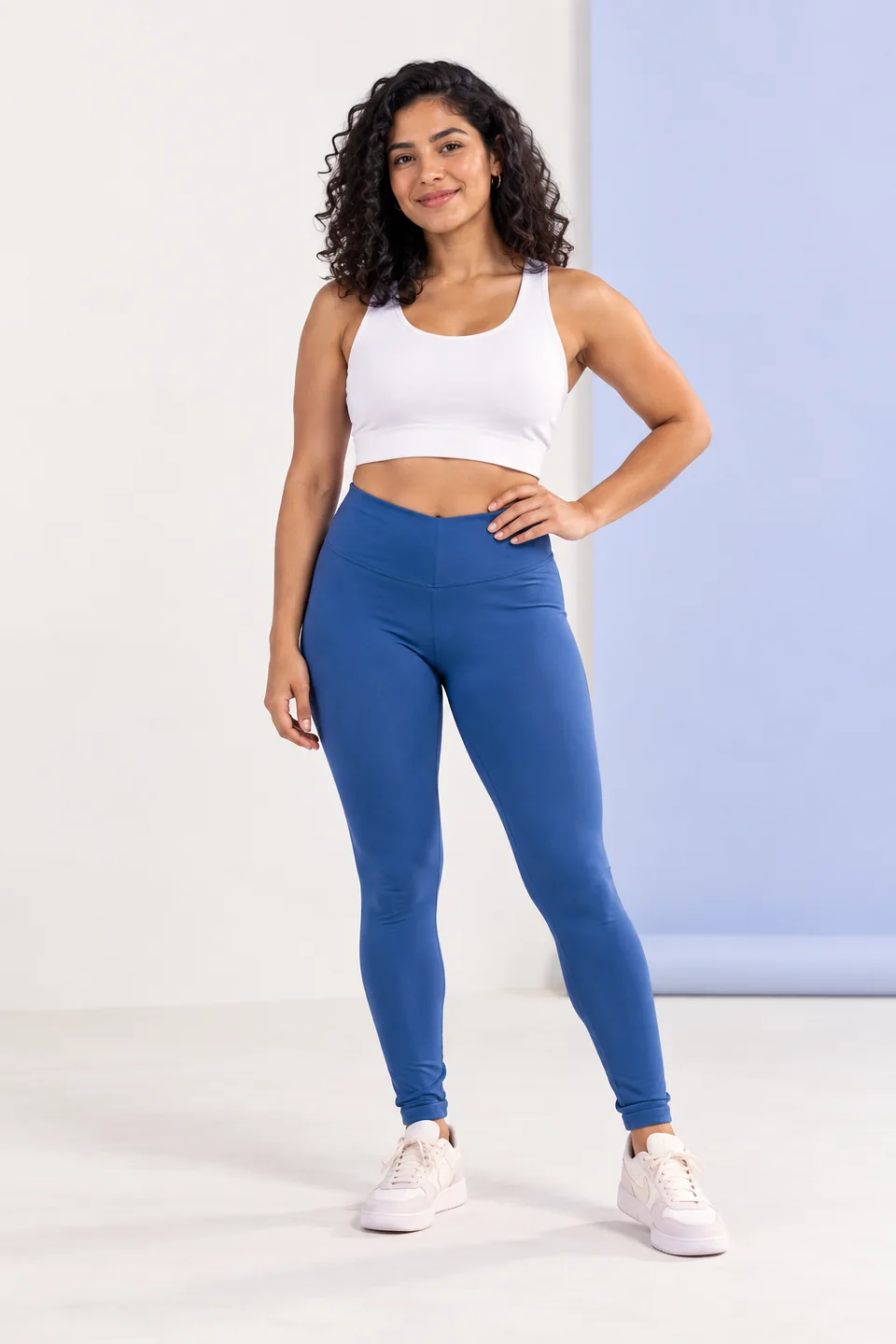Imagen editorial generada: mujer de cabello rizado con top blanco y leggings azules en un estudio claro.